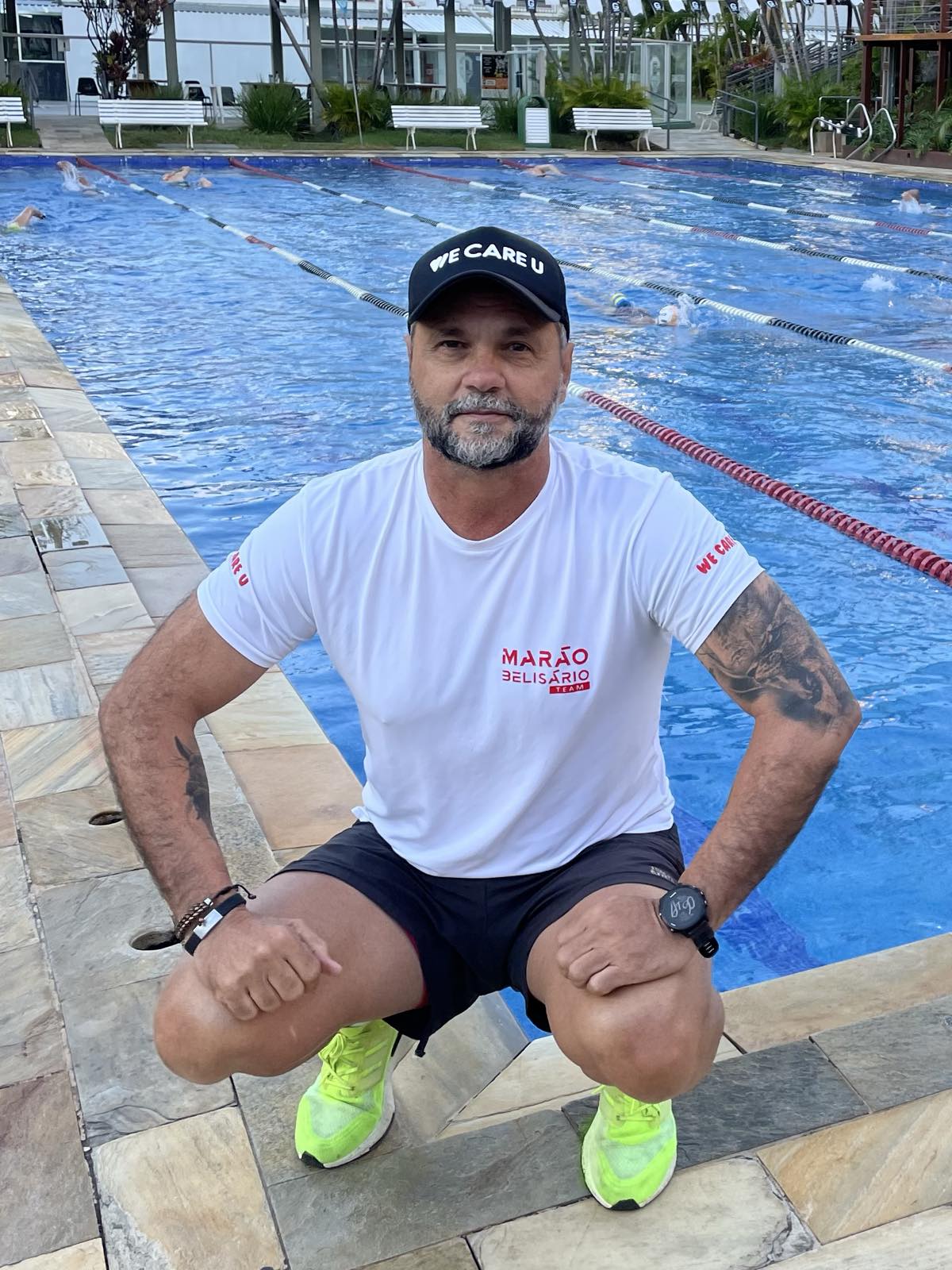 Marão Belisário agachado na borda da piscina, com atletas nadando nas raias ao fundo